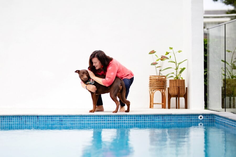 woman crouching down to hug a brown dog white background and reflection in blue pool